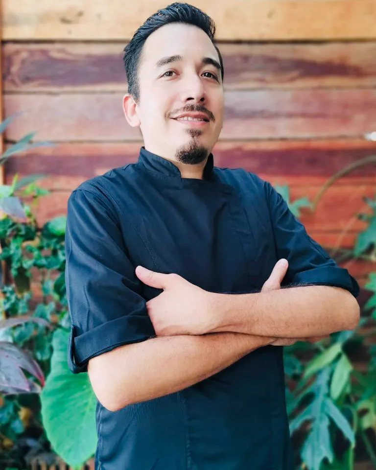 Chef Juan Francisco Quintero in a black chef's jacket, arms crossed, standing in front of tropical foliage